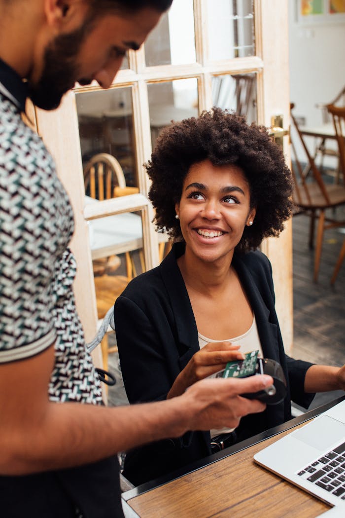 A young woman makes a contactless payment with a bank card at a café, smiling at the waiter.