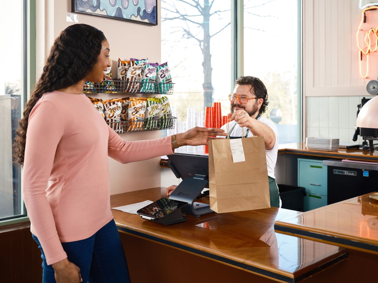 gallery-1 A customer receives a takeout order at a cafe counter, showcasing modern point of sale technology.
